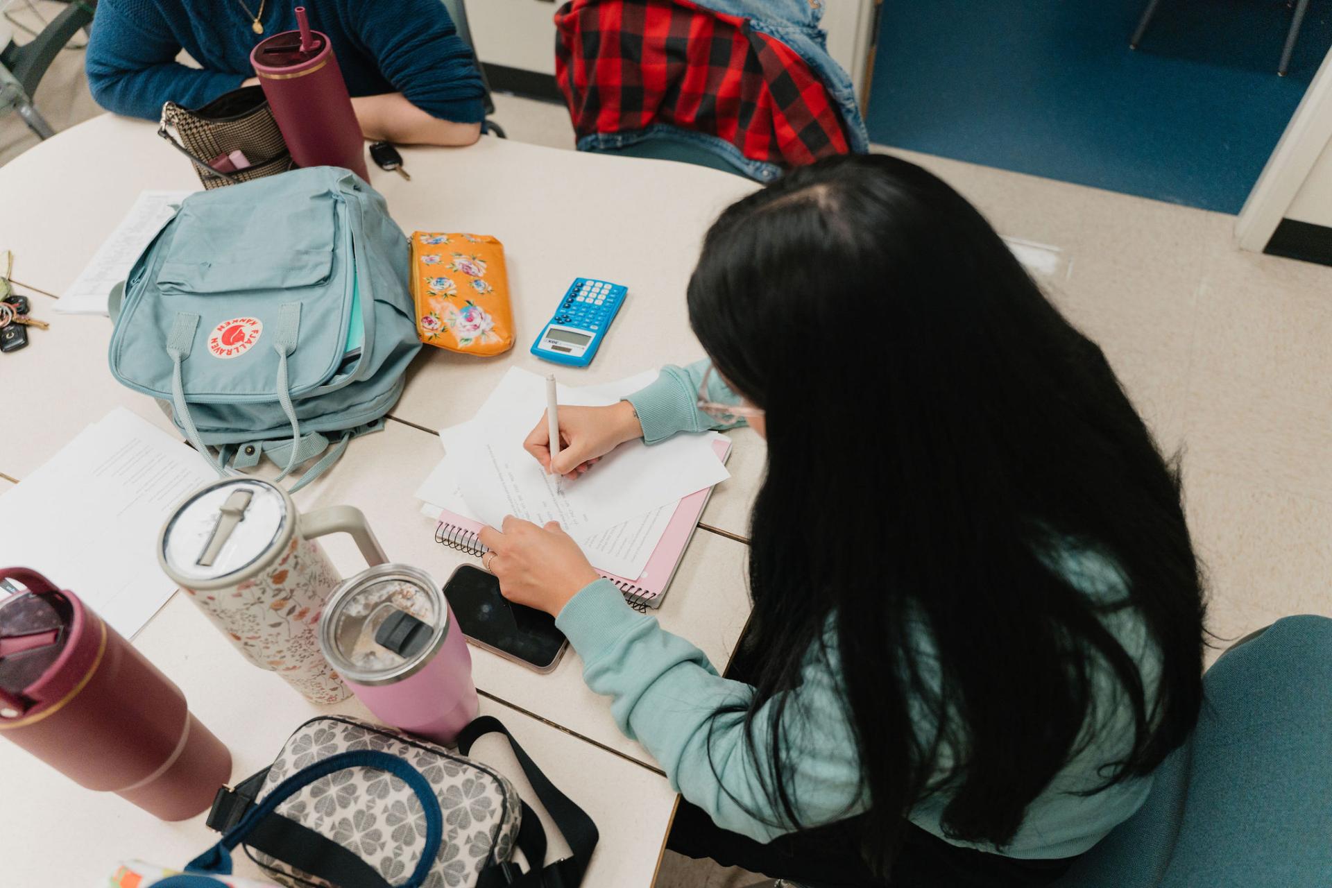 A female student with long, dark hark is taking notes at her desk with her phone, drinks, calculator, backpack and purse also on the desk. 