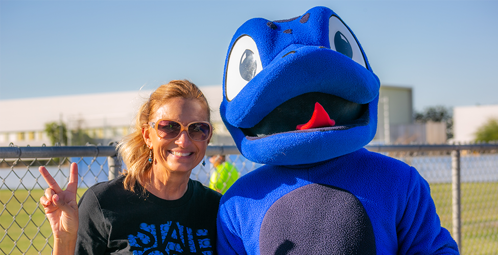 A woman wearing sunglasses smiles and holds up a peace sign while standing next to a large blue gecko mascot outdoors on a sunny day, with a chain-link fence and campus buildings in the background.