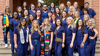 A large group of nursing graduates in navy blue scrubs stands together on outdoor steps in front of a brick building. Several students wear colorful stoles, cords, and pins, smiling proudly at the camera.