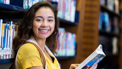 Female Hispanic student smiling at the camera with a book in the library