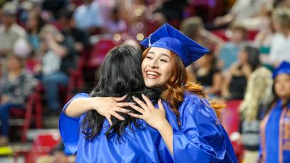A smiling female in a blue graduation cap and gown hugs another female graduate after the ceremony