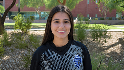 GateWay Community College student-athlete Lily Coronel smiles outdoors while wearing a black GateWay soccer jersey, standing in front of greenery and campus landscaping.