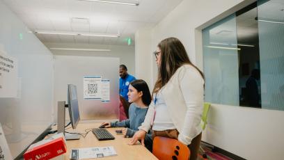 A female GateWay College employee in a white sweater stands beside a female student on a computer, helping with enrollment