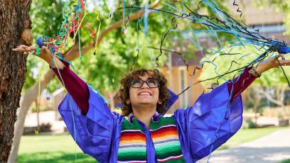 A student in a blue graduation cap and gown throws confetti and smiles