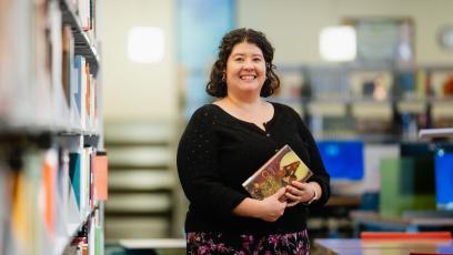 Amanda Takacs, GateWay Faculty Librarian, smiles wearing a black shirt and holding a book in the library