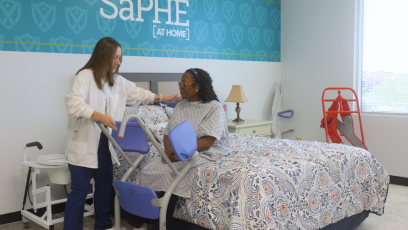 A female caregiver helps a female patient stand from a bed in the SaPHE lab at GateWay. 