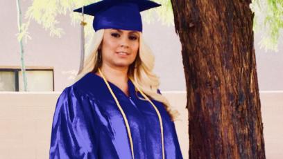 Female student standing outside next to tree, wearing blue cap and gown