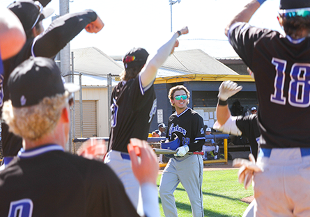 GateWay Geckos baseball player wearing sunglasses and holding a bat walks toward cheering teammates outside the dugout during a game on a sunny day.