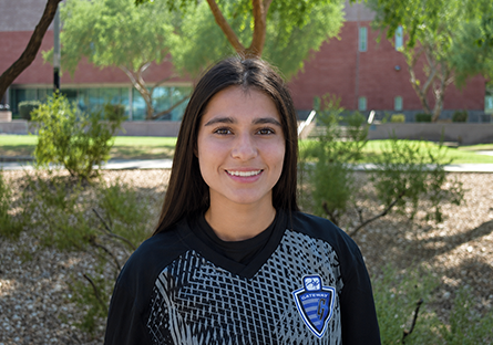 GateWay Community College student-athlete Lily Coronel smiles outdoors while wearing a black GateWay soccer jersey, standing in front of greenery and campus landscaping.