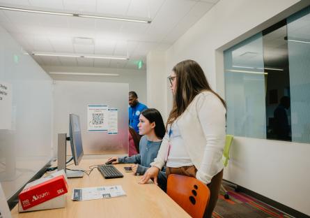 A female GateWay College employee in a white sweater stands beside a female student on a computer, helping with enrollment