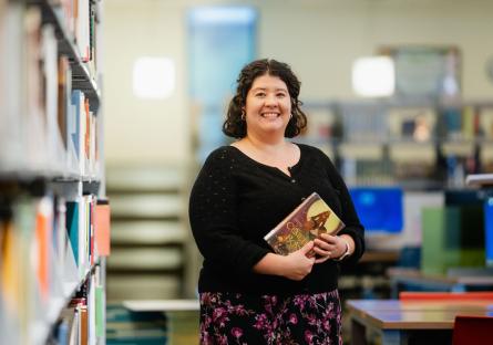 Amanda Takacs, GateWay Faculty Librarian, smiles wearing a black shirt and holding a book in the library