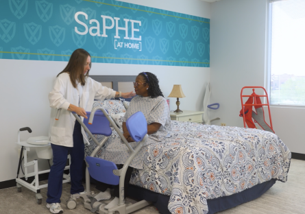 A female caregiver helps a female patient stand from a bed in the SaPHE lab at GateWay. 