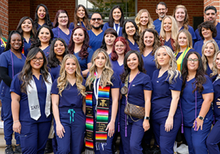 A large group of nursing graduates in navy blue scrubs stands together on outdoor steps in front of a brick building. Several students wear colorful stoles, cords, and pins, smiling proudly at the camera.