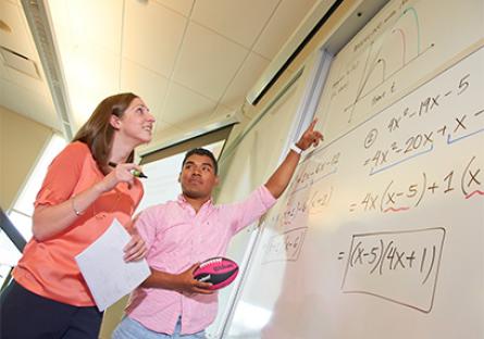 A teacher and student look at a math problem drawn up on a white board in a classroom