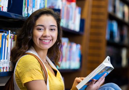 Female Hispanic student smiling at the camera with a book in the library