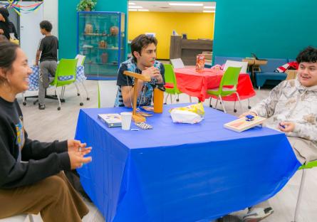 Juniper in a group of three people sitting at a table with a blue tablecloth, engaged in conversation in a colorful room.