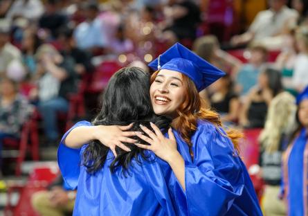 A smiling female in a blue graduation cap and gown hugs another female graduate after the ceremony