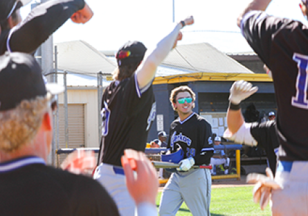 GateWay Geckos baseball player wearing sunglasses and holding a bat walks toward cheering teammates outside the dugout during a game on a sunny day.
