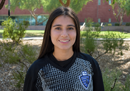 GateWay Community College student-athlete Lily Coronel smiles outdoors while wearing a black GateWay soccer jersey, standing in front of greenery and campus landscaping.