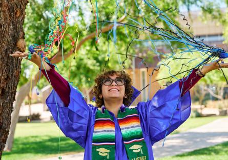 A student in a blue graduation cap and gown throws confetti and smiles
