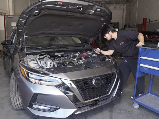 Student look under engine of silver car