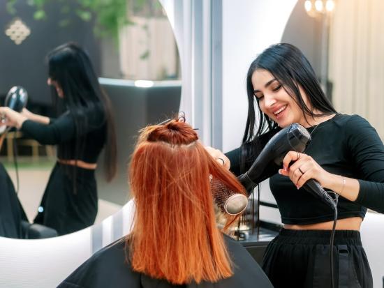 Professional hairstylist blow-drying and styling a client's red hair in a salon setting.