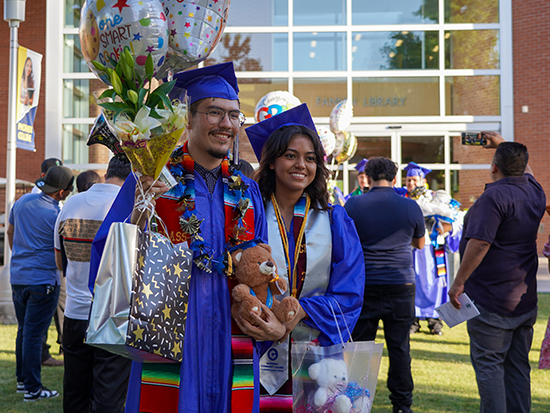 Photo a male and female high school students at their graduation celebration. They are in a blue cap and gown and adorned with tassels and stoles. 