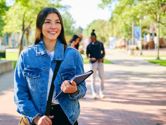 A smiling college student wearing a denim jacket and carrying a notebook walks on a sunny campus pathway.