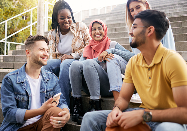 Group of five diverse young adults sitting on outdoor steps, smiling and talking together in a relaxed, friendly setting.