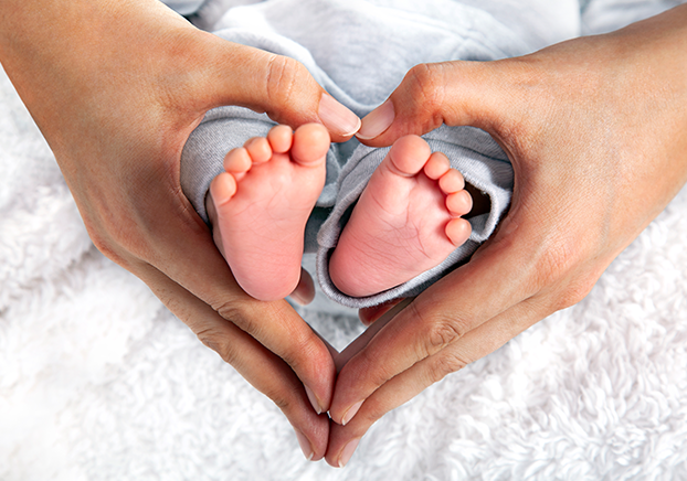 Close-up of adult hands forming a heart shape around a newborn’s feet, resting on a soft white blanket.