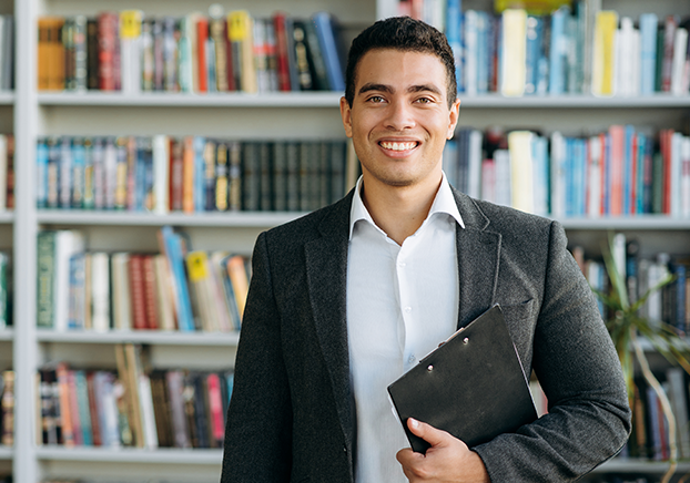 Young man in a suit jacket holding a clipboard, standing in front of a bookshelf in a library.