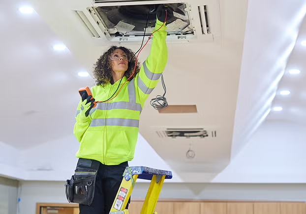 Woman standing on ladder checking air conditioning unit in ceiling.