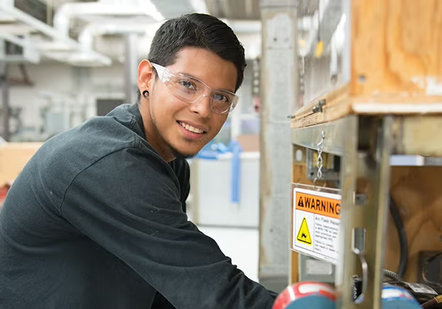 Male student working on air conditioning unit smiling at camera.