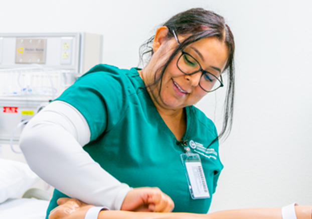 A woman in green medical scrubs and glasses practices a clinical skill on a mannequin arm, smiling as she works in a healthcare training lab.