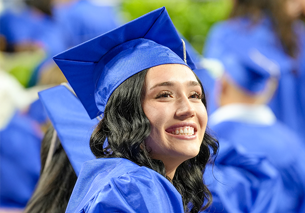 smiling graduate in a bright blue cap and gown looks over her shoulder during a commencement ceremony. Other graduates in matching regalia are seated in the background.