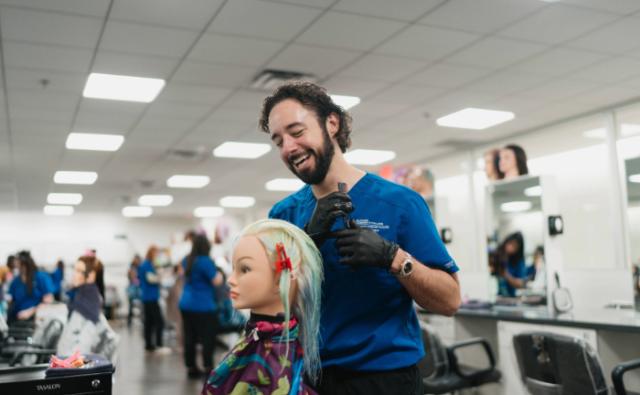 Male cosmetology student in blue scrubs happily practicing hair styling on a mannequin in a campus salon